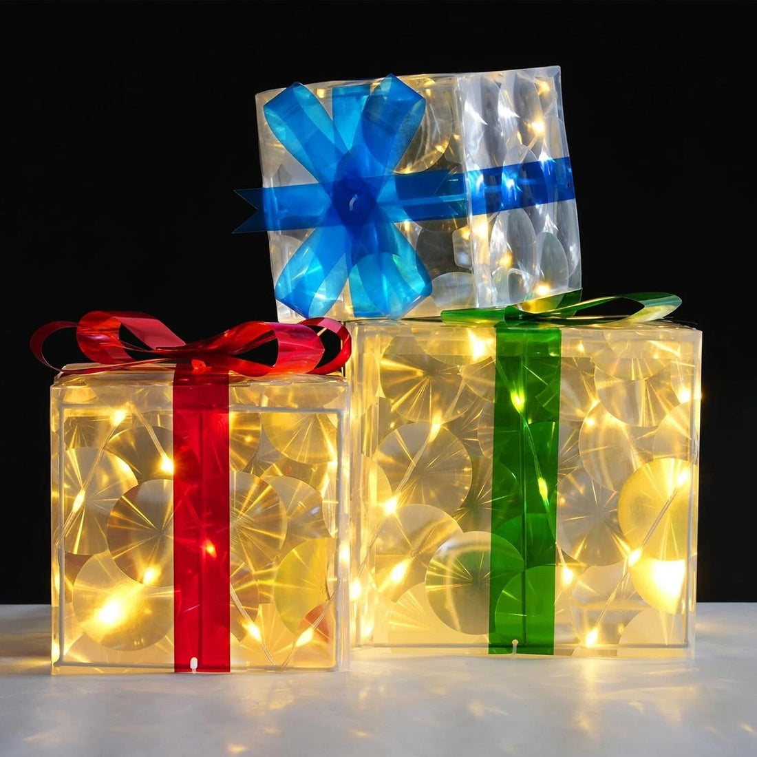Three lit Christmas gift boxes in small, medium, and large sizes with red, green, and blue bows glowing with warm LED lights