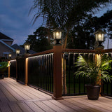 Two matte black solar post cap lights with glass shades mounted on 4x4 wood fence posts glowing warm white LED light - view 2